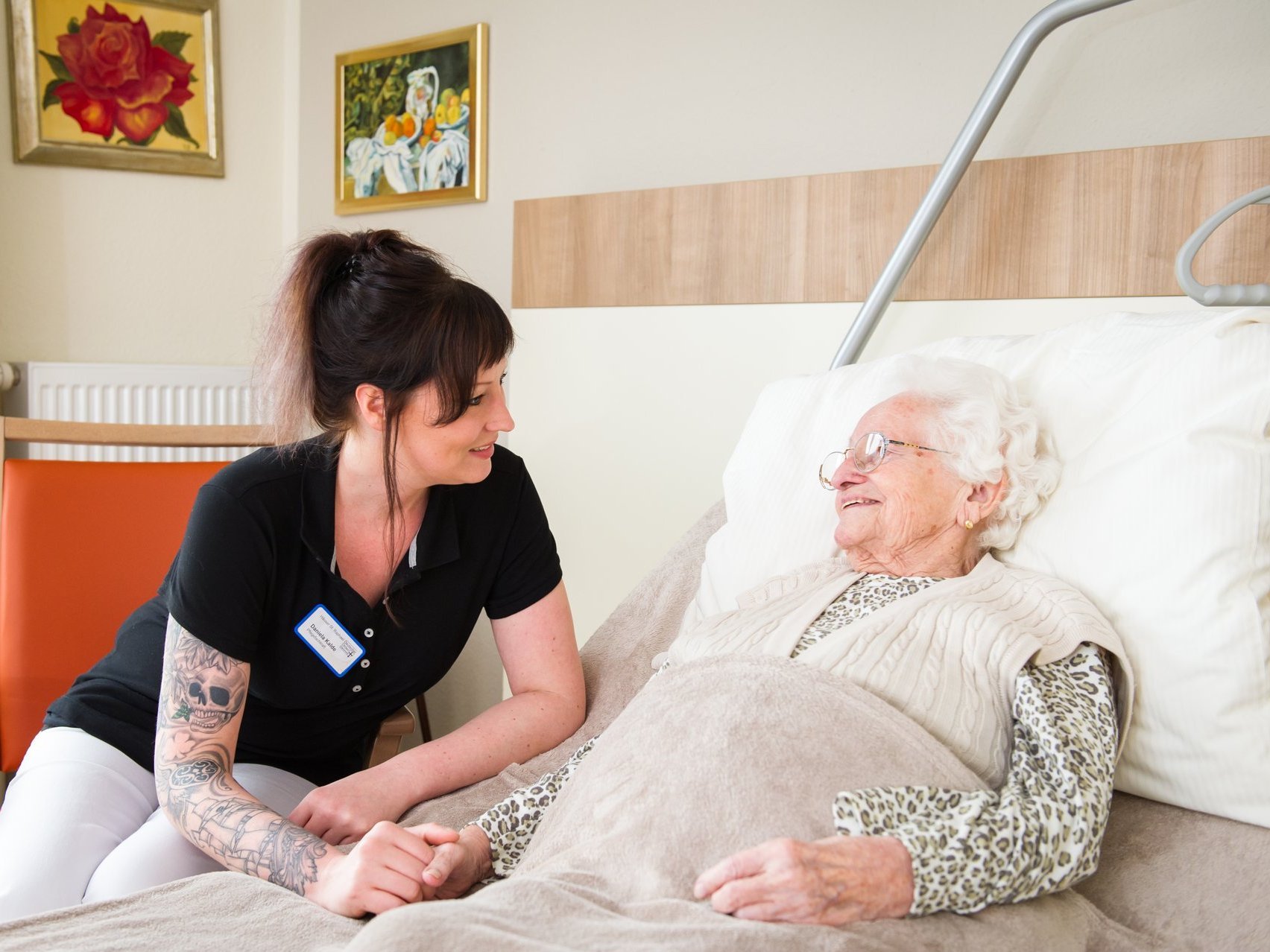 Eine junge Pflegerin kümmert sich um eine weisshaarige Frau, die in einem Krankenbett liegt - sie halten Hand und lächeln sich an