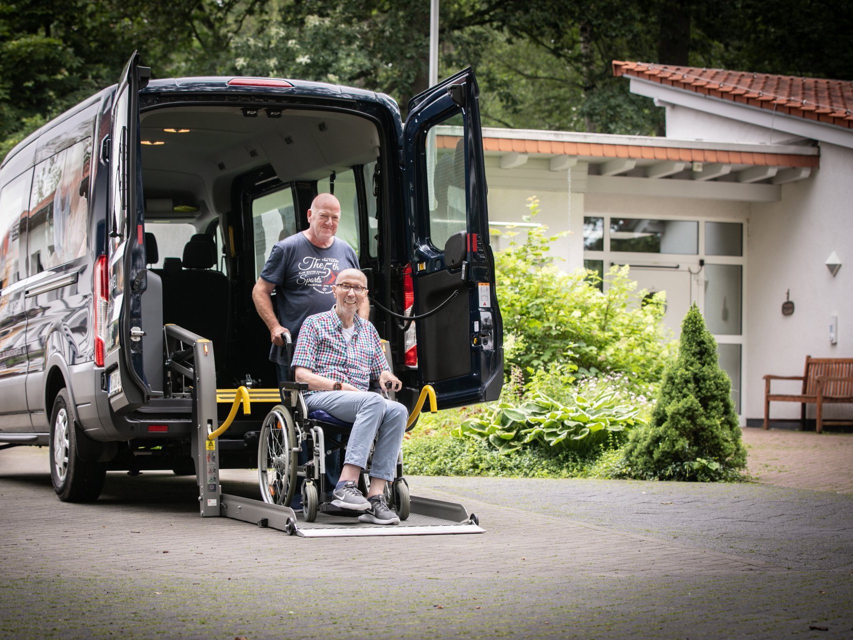 Ein Mitarbeiter vom Fahrdienst steht vor einem schwarzen Transporter und schiebt einen Bewohner im Rollstuhl.