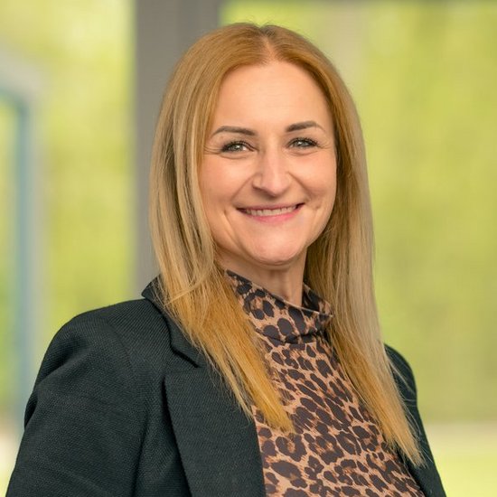 Anna Schulte Porträt einer lächelnden Frau mit langen blonden Haaren. Sie trägt einen schwarzen Blazer über einem Leopardenprint-Oberteil und Jeans und steht vor einem unscharfen Fensterhintergrund.