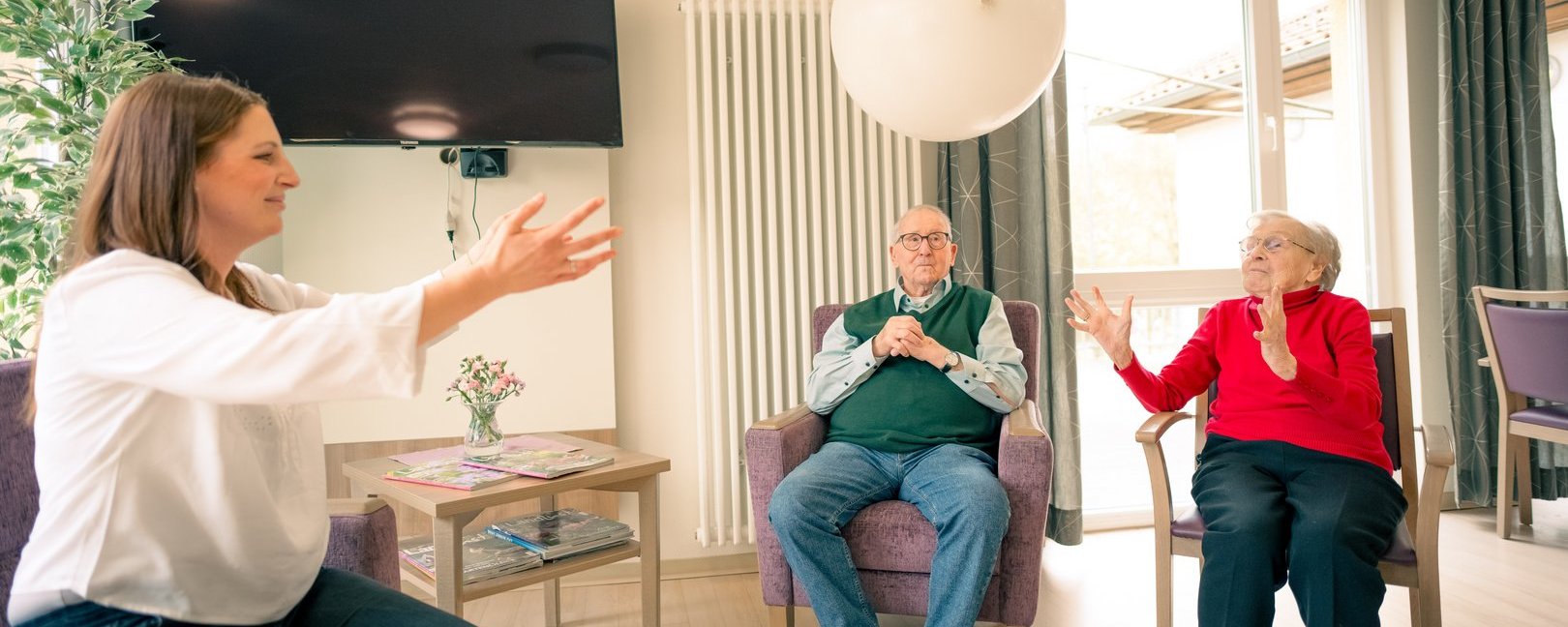 Eine junge Frau und zwei Senioren spielen in einem hellen Raum. Die Frau links und die ältere Dame rechts werfen sich einen weißen Ballon zu, während ein älterer Herr in der Mitte zuschaut.