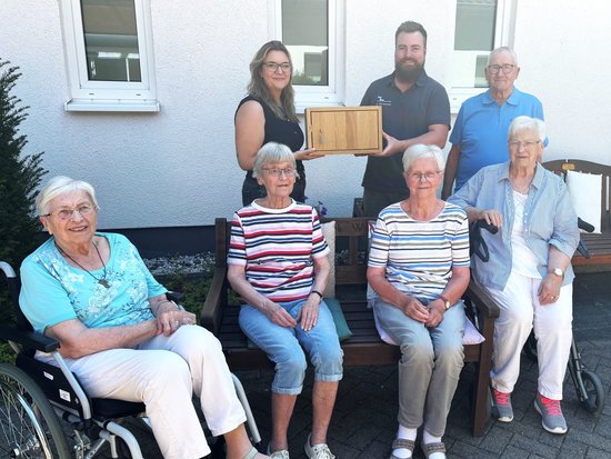 Vor einem weißen Gebäude posieren sieben Personen für ein Foto. Fünf ältere Frauen sitzen im Vordergrund, wobei die linke Frau einen Rollstuhl nutzt. Dahinter stehen drei Personen, ein Mann in der Mitte hält eine Holzplakette. Die Szene deutet auf eine feierliche Übergabe oder ein Gruppenfoto hin.