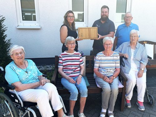 Vor einem weißen Gebäude posieren sieben Personen für ein Foto. Fünf ältere Frauen sitzen im Vordergrund, wobei die linke Frau einen Rollstuhl nutzt. Dahinter stehen drei Personen, ein Mann in der Mitte hält eine Holzplakette. Die Szene deutet auf eine feierliche Übergabe oder ein Gruppenfoto hin.