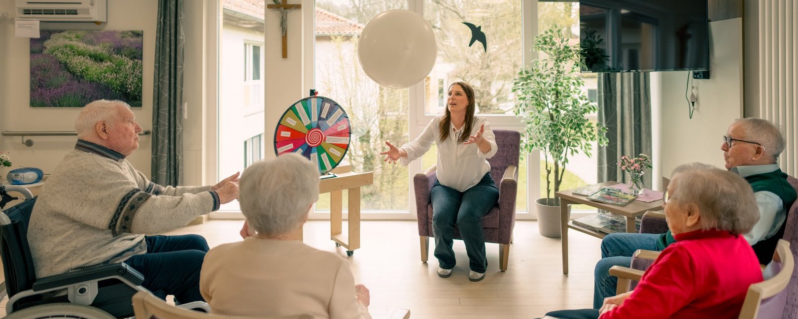 Eine Betreuerin führt vier Senioren in einem hellen Raum an. Einer sitzt im Rollstuhl. Auf einem Tisch steht ein buntes Glücksrad. Die älteren Menschen sitzen im Kreis und schauen aufmerksam zur Betreuerin, die einen Ballon hin und her wirft.