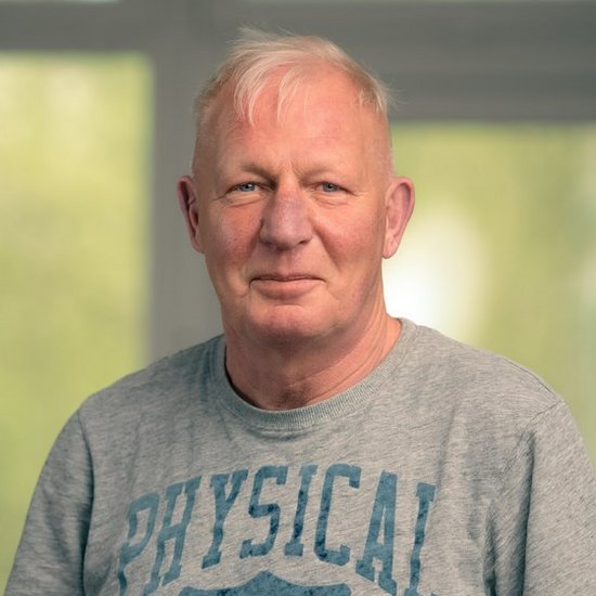 Andreas Bahne Ein Porträt eines älteren Mannes mit kurzen weißen Haaren. Er steht frontal zur Kamera, die Hände in den Hosentaschen, und trägt ein graues T-Shirt mit dem blauen Schriftzug „PHYSICAL EDUCATION". Der Hintergrund zeigt ein unscharfes Fenster mit Blick ins Grüne.