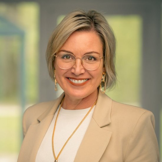 Sandra Duken Porträt einer lächelnden Frau mit kurzen blonden Haaren und Brille. Sie trägt einen beigen Blazer, ein weißes Top und eine lange Halskette. Der Hintergrund ist unscharf.