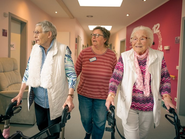 Zwei Seniorinnen gehen mit Rollatoren durch einen hellen Flur, begleitet von einer Pflegerin. Die Betreuerin läuft zwischen den Frauen und unterstützt sie beim Gehen, was eine Szene von Fürsorge und aktiver Mobilität darstellt.