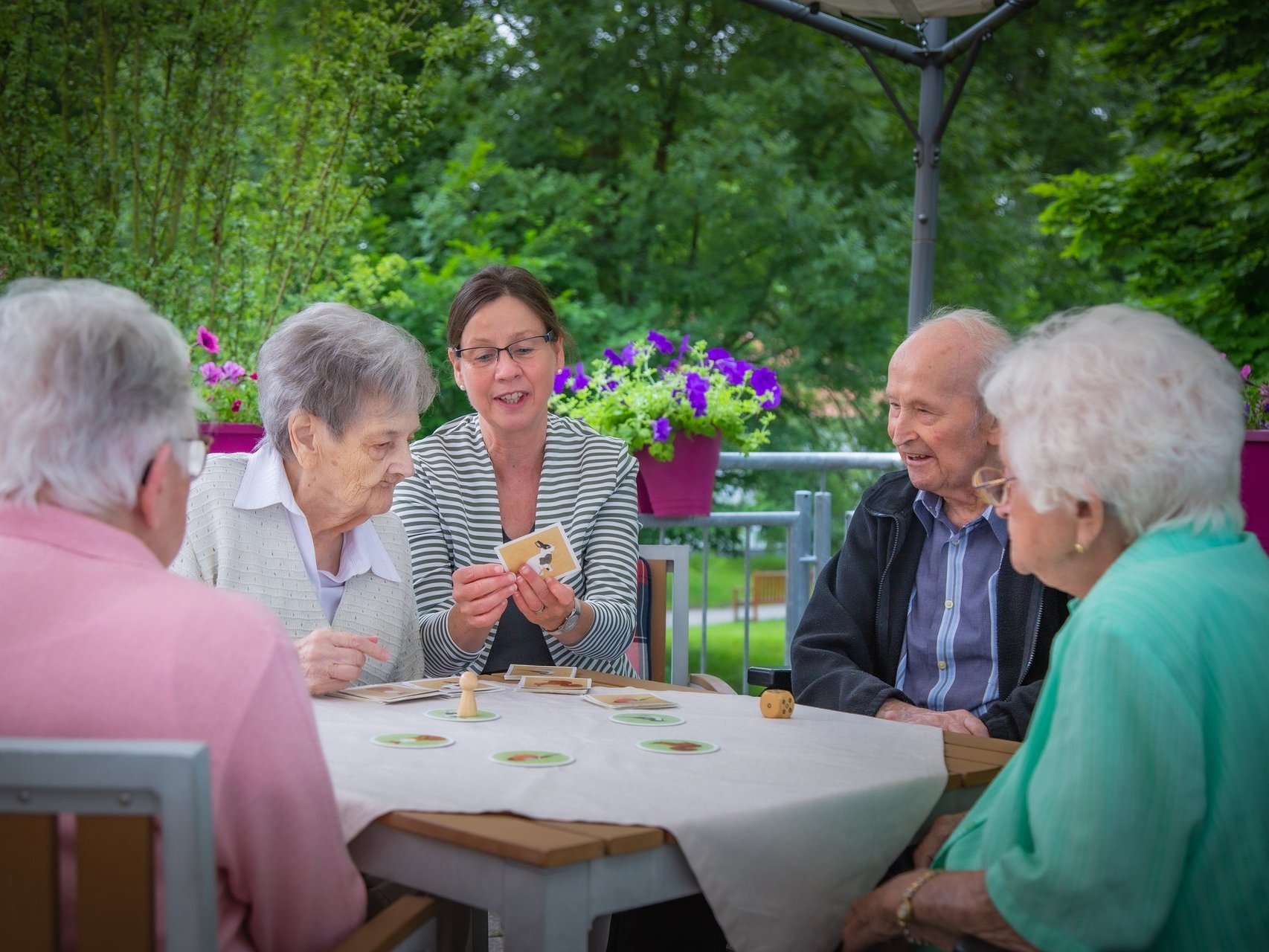 Vier Bewohner spielen mit einer Mitarbeiterin ein Brettspiel auf einer Terasse im Garten