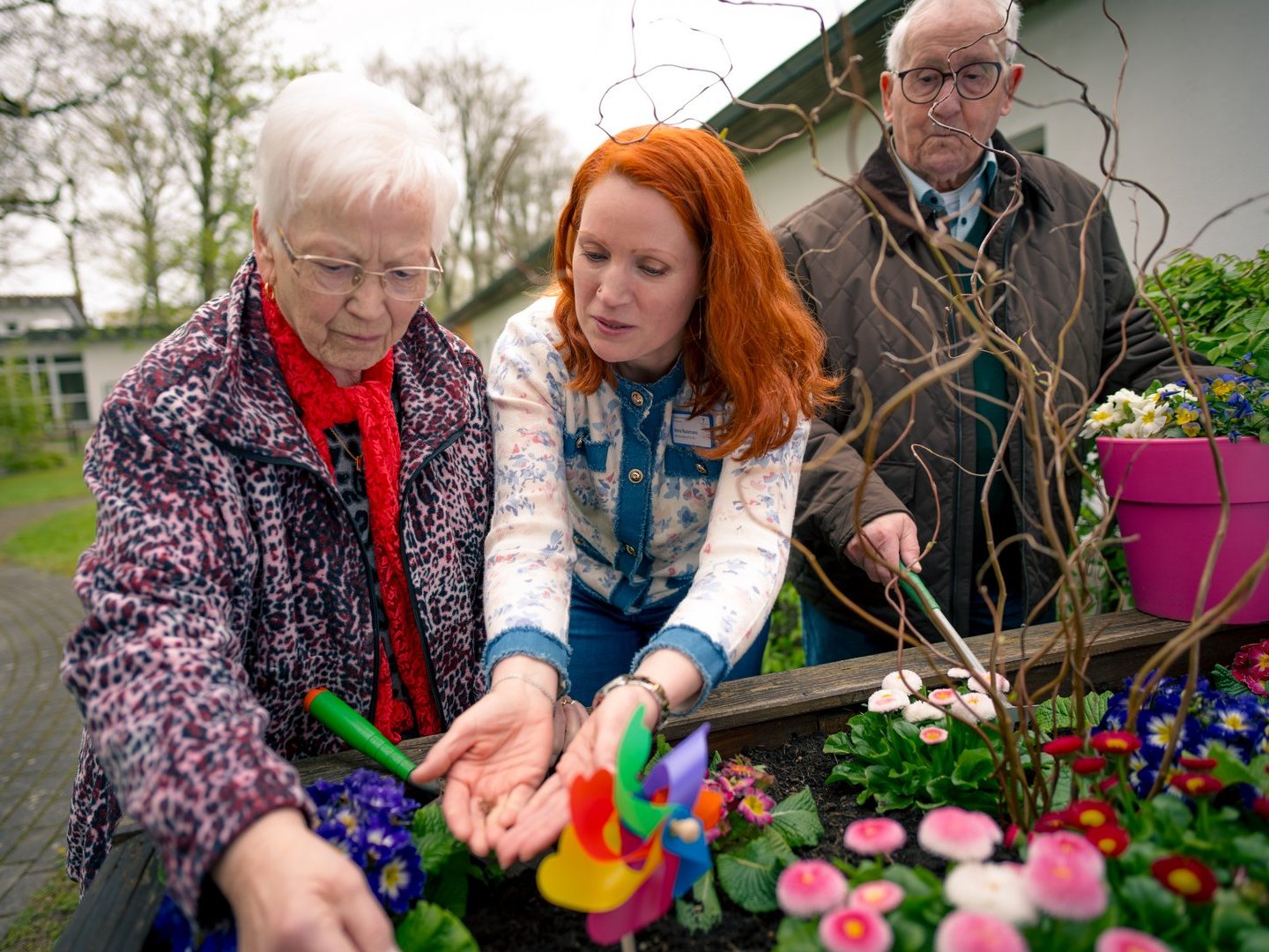Eine ältere Dame und eine jüngere Frau mit roten Haaren arbeiten gemeinsam an einem Hochbeet. Die jüngere Frau hält ein buntes Windrad, die ältere Frau berührt die Pflanzen. Im Hintergrund steht ein älterer Mann mit einer Schaufel.