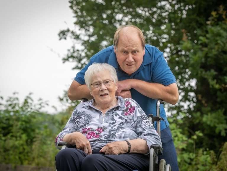 Eine Frau sitzt im Rollstuhl - der Sohn stützt sich für das Foto auf die Griffe des selbigen