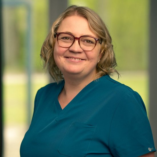 Tatjana Moor Eine lächelnde Frau mit blonden Haaren und Brille trägt blaue medizinische Kleidung und steht vor einem unscharfen Fenster.