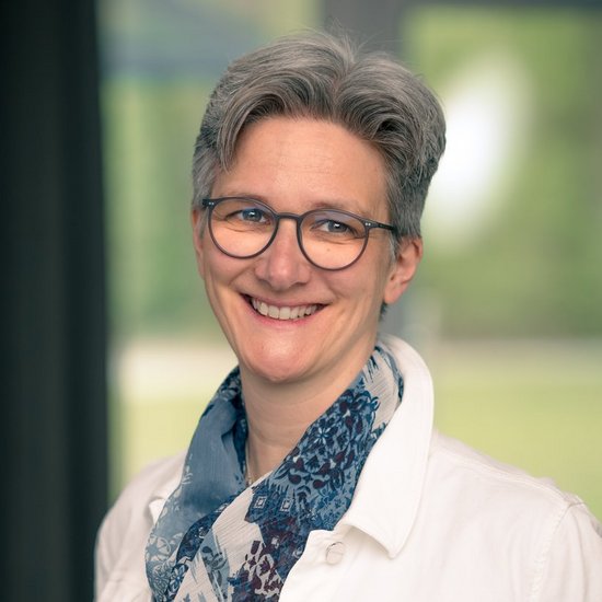 Margit Bartmann Ein Porträt einer lächelnden Frau mit kurzen grauen Haaren und Brille. Sie trägt eine weiße Jacke und einen blau gemusterten Schal vor einem unscharfen Fensterhintergrund.
