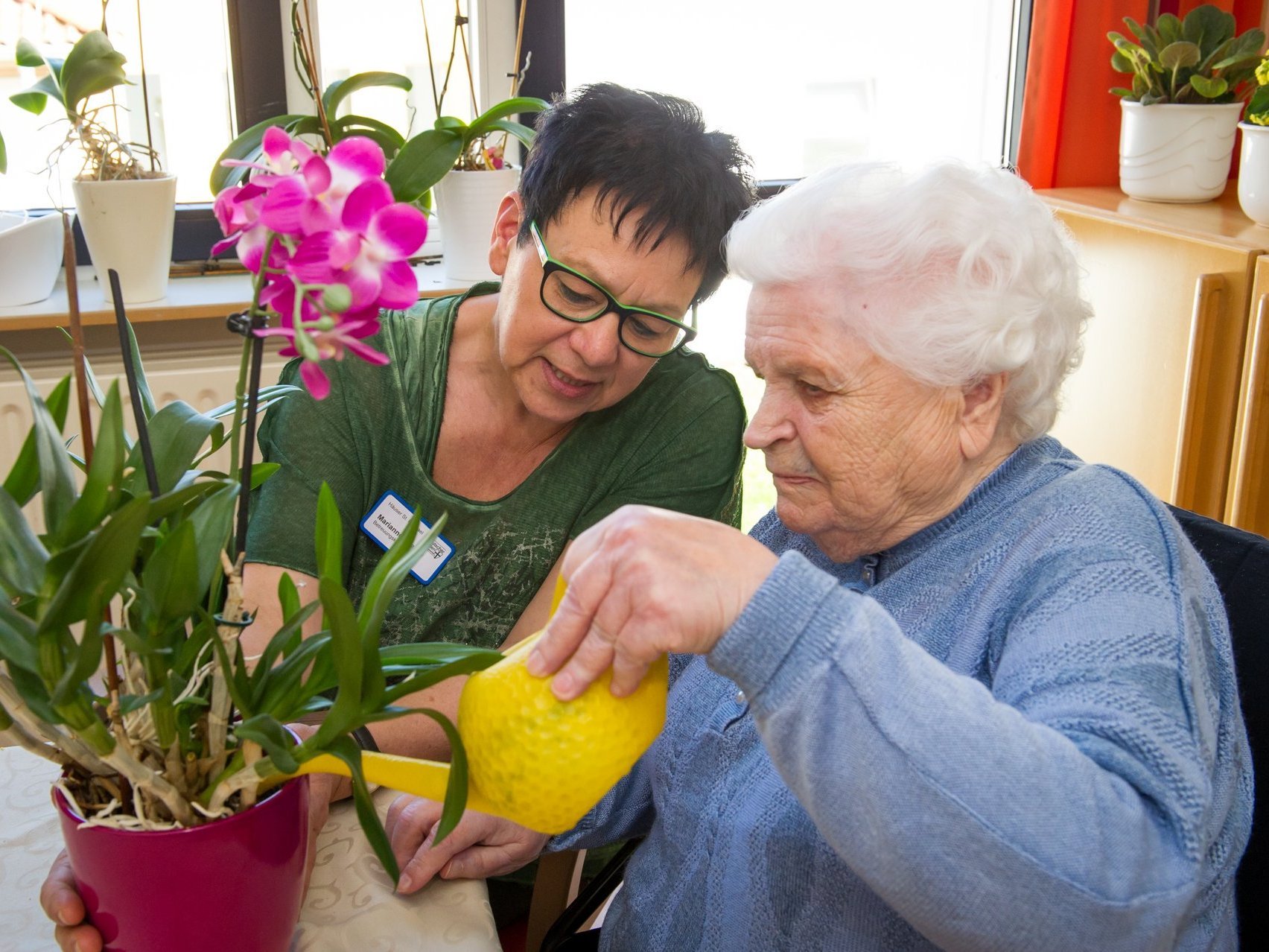 Eine Mitarbeiterin hilf einer Heimbewohnerin beim Blumengießen mit einer gelben Gießkanne.