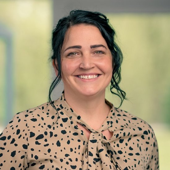 Katja Henschke Ein Porträt einer lächelnden Frau mit dunklen Haaren. Sie trägt eine beige Bluse mit schwarzen Flecken und steht vor einem unscharfen Fenster mit Blick ins Grüne.