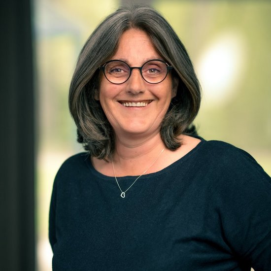 Angela Junghölter Eine lächelnde Frau mit schulterlangen, grau melierten Haaren und einer runden Brille blickt freundlich in die Kamera. Sie trägt ein dunkles Oberteil und eine feine Halskette vor einem hellen, unscharfen Hintergrund.