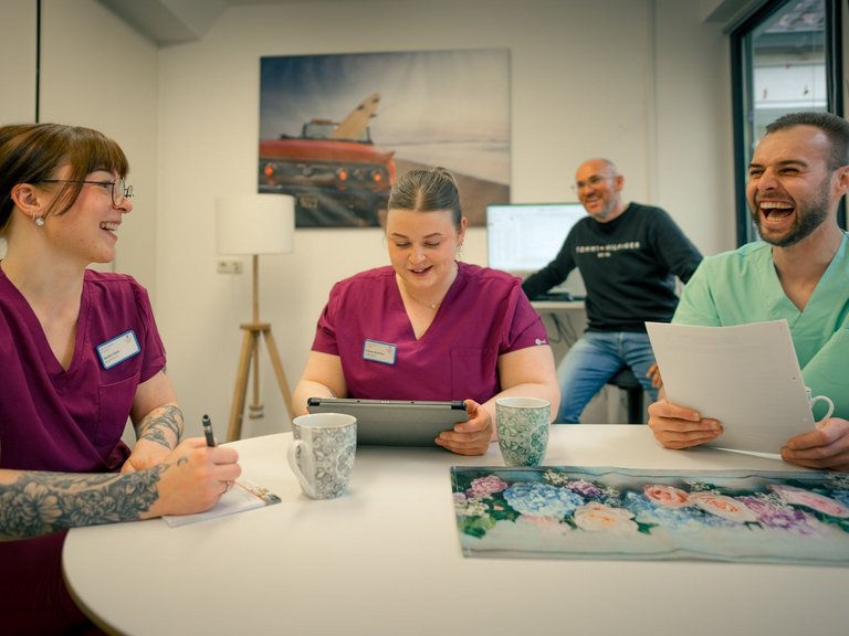 Drei Personen in medizinischen Kitteln sitzen an einem Tisch und lachen. Eine Frau schreibt, eine andere nutzt ein Tablet, und ein Mann hält Papiere. Im Hintergrund steht ein weiterer Mann und lächelt. Die Szene zeigt eine fröhliche Team-Pause.