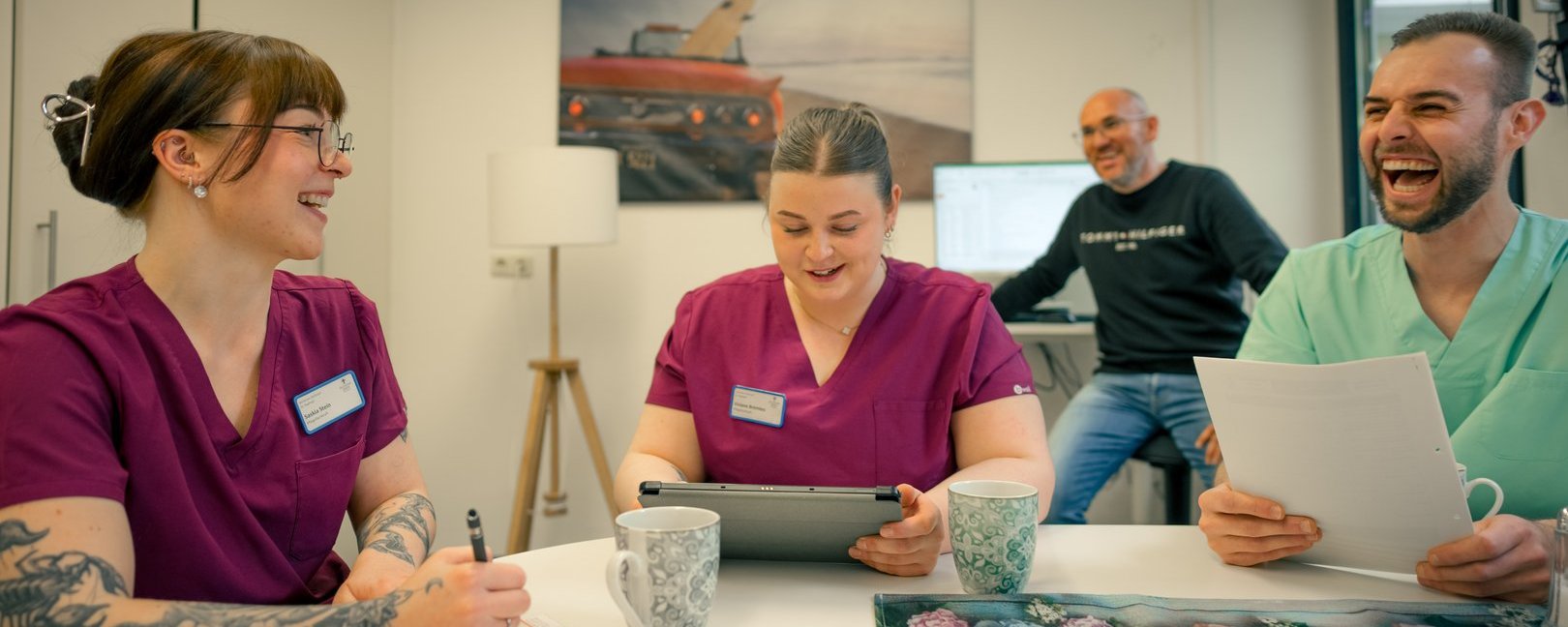 Drei Personen in medizinischen Kitteln sitzen an einem Tisch und lachen. Eine Frau schreibt, eine andere nutzt ein Tablet, und ein Mann hält Papiere. Im Hintergrund steht ein weiterer Mann und lächelt. Die Szene zeigt eine fröhliche Team-Pause.