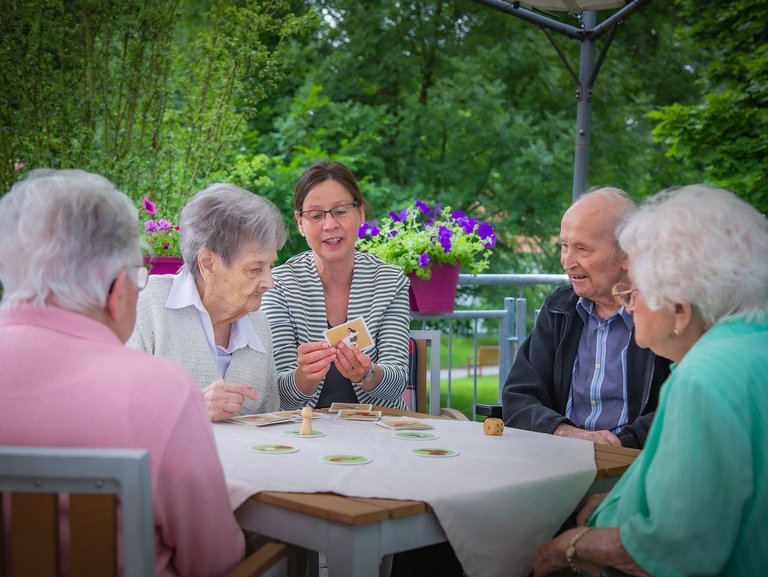 Eine Pflegerin unterstützt Bewohner/innen beim Kartenspiel auf der Terasse