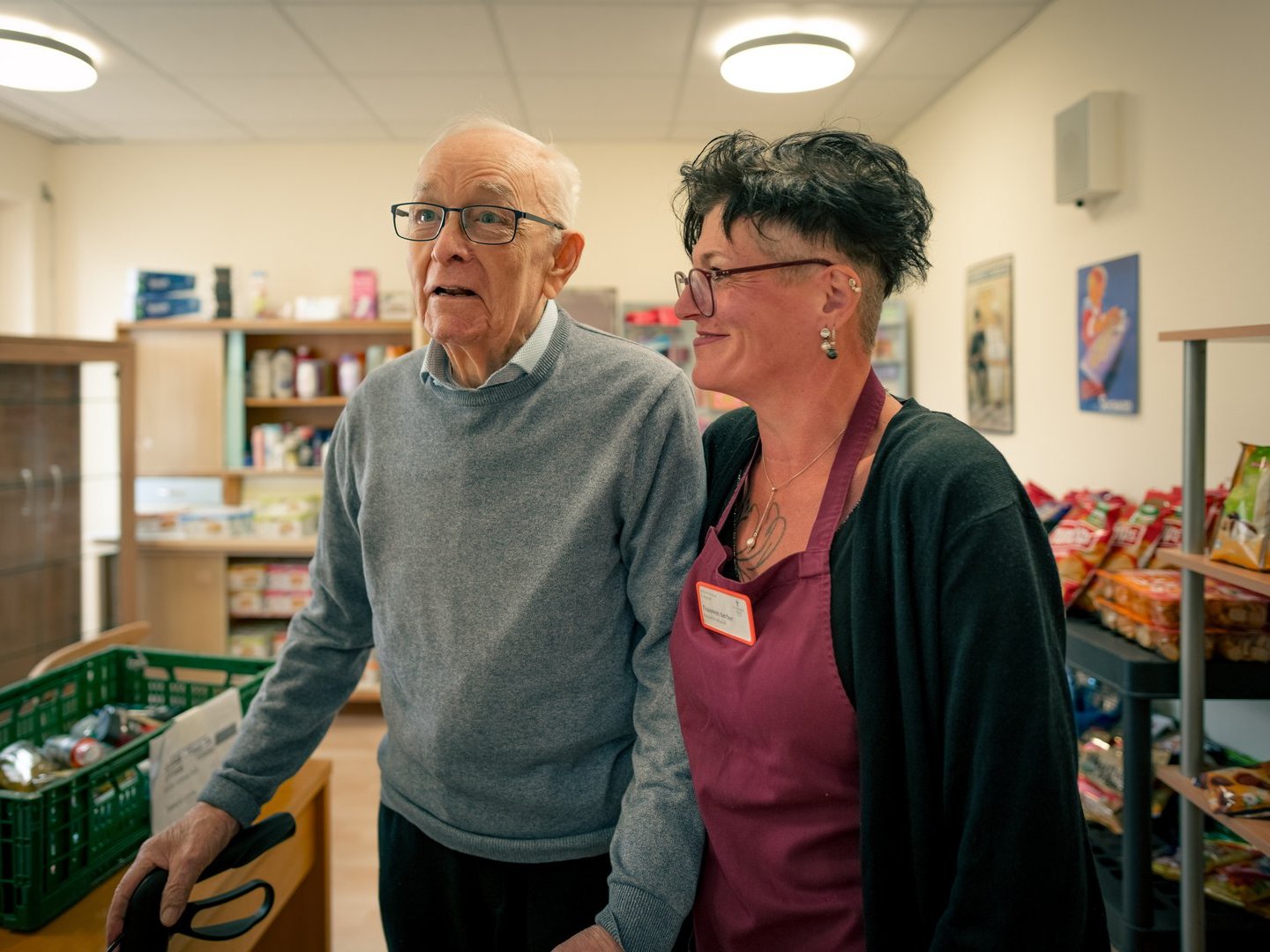 Ein älterer Mann und eine Frau in einer Schürze stehen gemeinsam in einem kleinen Laden. Der Mann hält sich an seinem Rollator fest, während beide in dieselbe Richtung blicken. Im Hintergrund sind Regale mit Waren zu sehen.