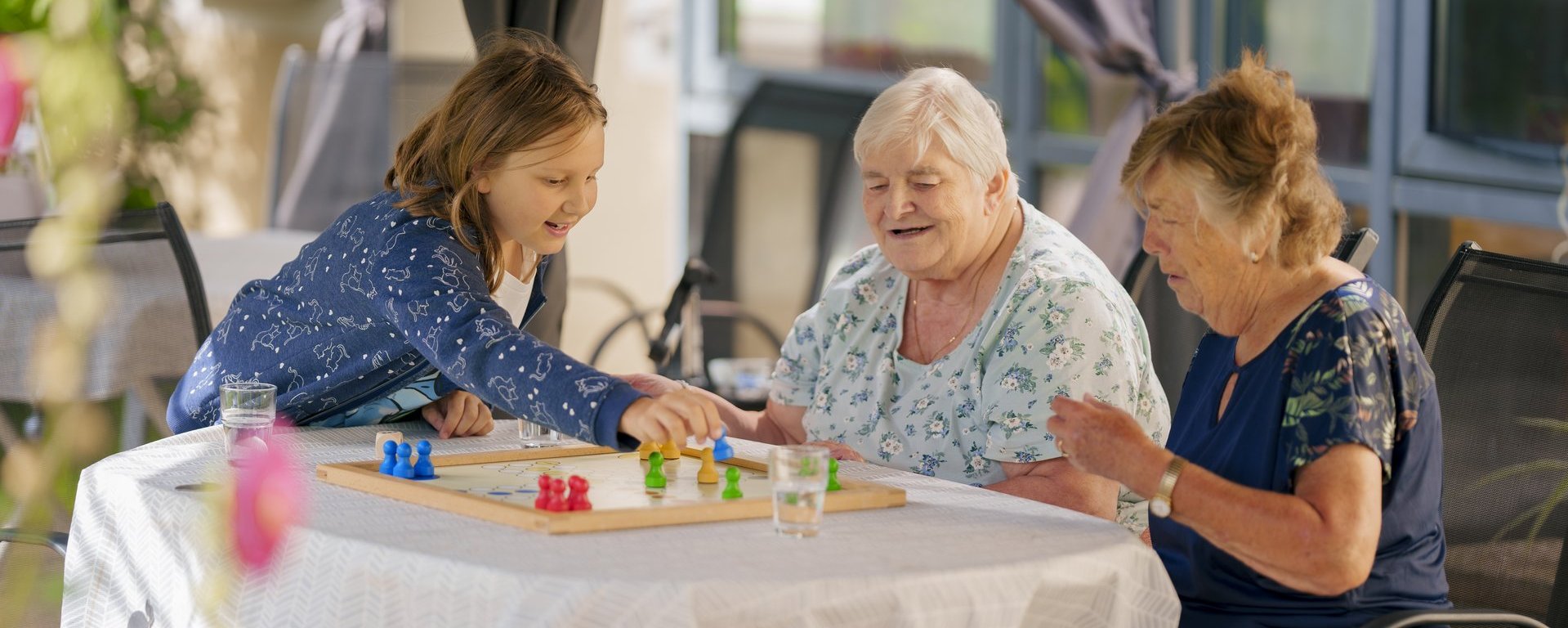 Ein junges Mädchen und zwei ältere Frauen spielen fröhlich ein Brettspiel im Freien. Sie sitzen an einem Tisch und lachen gemeinsam, was eine warme, generationenübergreifende Atmosphäre zeigt.