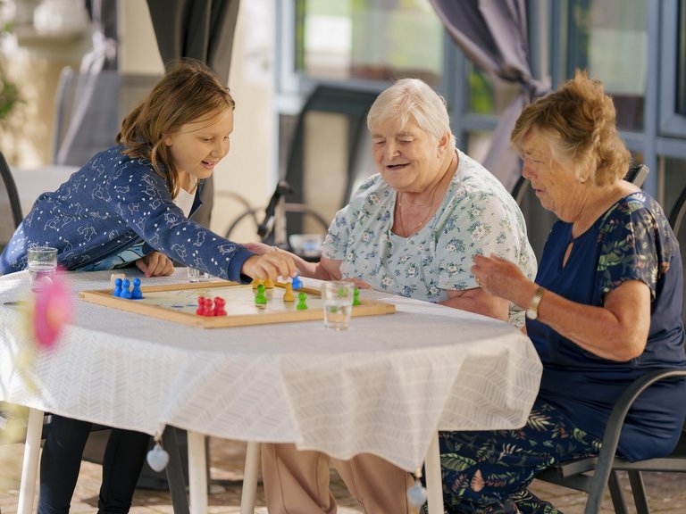 Ein junges Mädchen und zwei ältere Frauen spielen fröhlich ein Brettspiel im Freien. Sie sitzen an einem Tisch und lachen gemeinsam, was eine warme, generationenübergreifende Atmosphäre zeigt.