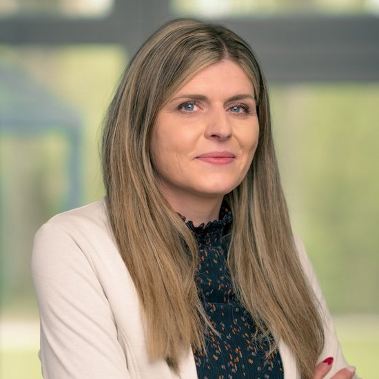 Patrycja Smietana Porträt einer Frau mit langen blonden Haaren. Sie trägt einen hellen Blazer über einer dunklen, gemusterten Bluse und hat die Arme verschränkt. Ihr Blick ist leicht zur Seite gerichtet vor einem unscharfen Fensterhintergrund.
