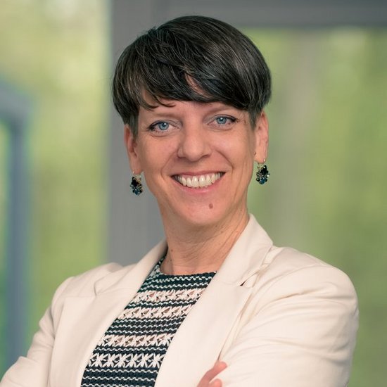 Julia Harnischmacher Ein Porträt einer lächelnden Frau mit kurzen dunklen Haaren. Sie steht selbstbewusst mit verschränkten Armen da und trägt einen cremefarbenen Blazer über einem schwarz-weiß gemusterten Oberteil. Im unscharfen Hintergrund ist ein Fenster mit Blick auf grüne Vegetation zu sehen.