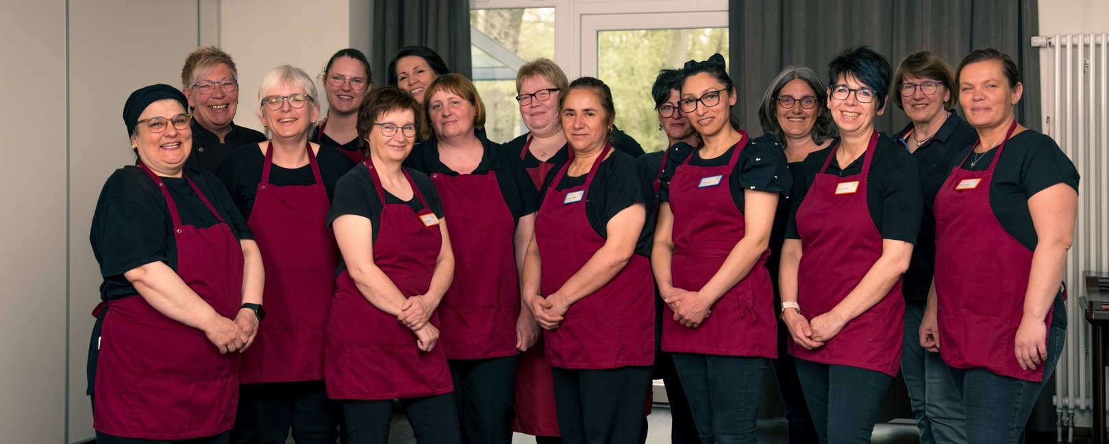 Ein Gruppenfoto von etwa 15 Mitarbeitern, überwiegend Frauen, die in einer Reihe stehen. Sie tragen einheitliche Arbeitskleidung mit schwarzen T-Shirts und dunkelroten Schürzen und lächeln in die Kamera.