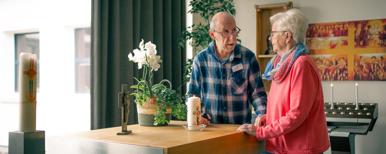Ein älterer Mann und eine ältere Frau steht vor einem Altar in einer Kirche und unterhält sich. Der Mann trägt ein kariertes Hemd, die Frau eine rosa Jacke. Auf dem Altar stehen Kerzen und ein Kreuz.