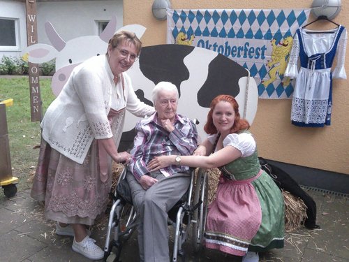 Drei Personen in bayerischer Tracht posieren vor einer Oktoberfest-Dekoration. Ein älterer Mann im Rollstuhl sitzt in der Mitte, flankiert von zwei Frauen, die ihn umarmen. Im Hintergrund sind ein Schild mit „Oktoberfest“, eine Kuh und ein Dirndl zu sehen.