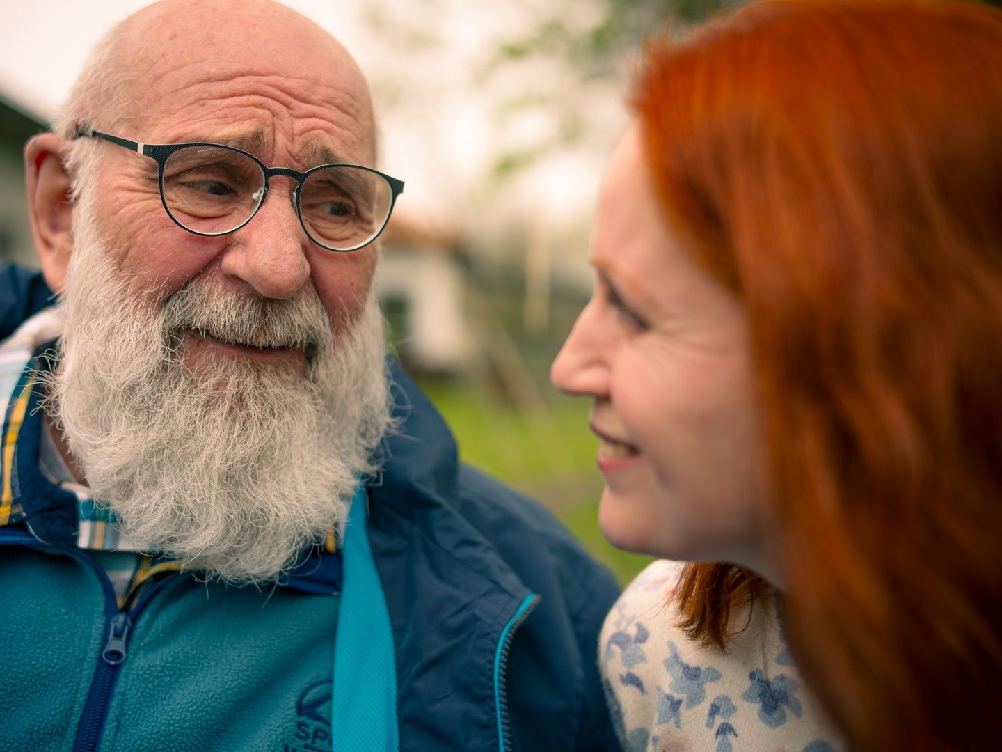 Ein älterer Mann mit weißem Bart und Brille lächelt eine junge Frau mit roten Haaren an. Beide blicken sich im Freien an und wirken glücklich.
