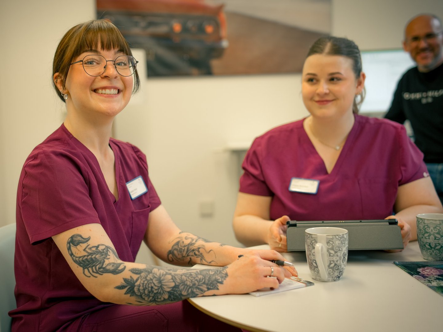 Zwei freundliche Mitarbeiterinnen in lila medizinischen Kitteln sitzen an einem Tisch und lächeln. Die Frau im Vordergrund hat Tattoos am Arm und hält einen Stift, während im Hintergrund ein Kollege steht.
