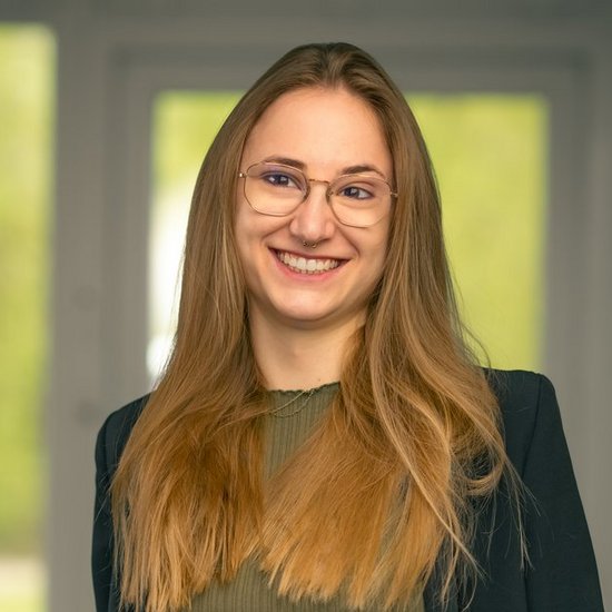 Louisa Rohe Eine lächelnde Frau mit langen blonden Haaren und Brille steht frontal vor einem unscharfen Fenster. Sie trägt ein olivgrünes Oberteil unter einer dunklen Jacke und hat die Hände locker in den Hosentaschen.
