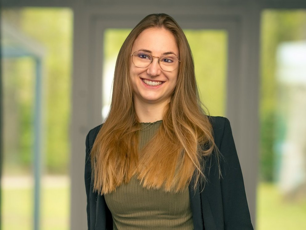 Eine lächelnde Frau mit langen blonden Haaren und Brille steht frontal vor einem unscharfen Fenster. Sie trägt ein olivgrünes Oberteil unter einer dunklen Jacke und hat die Hände locker in den Hosentaschen.