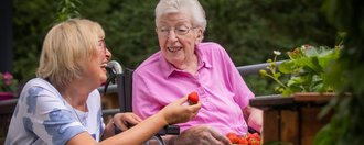 Eine Bewohnerin im Rollstuhl sitzt mit eirner Schale Erdbeeren auf dem Schoß im Garten und neben ihr kniet eine Besucherin und isst Erdbeeren.