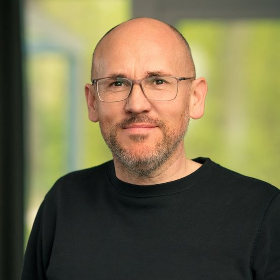 Christoph Ceiler Porträt eines kahlköpfigen Mannes mit Brille und Bart, der ein schwarzes Oberteil trägt. Er blickt direkt in die Kamera vor einem unscharfen Hintergrund.