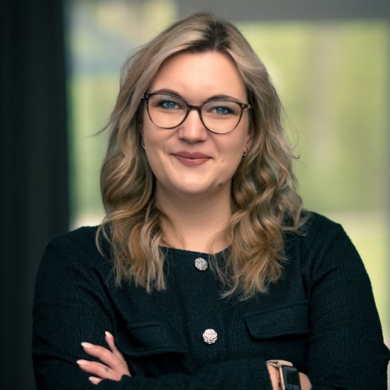 Laura Hesse Porträt einer lächelnden Frau mit blonden, welligen Haaren und einer Brille. Sie trägt eine dunkle Jacke und hat die Arme vor dem Körper verschränkt.