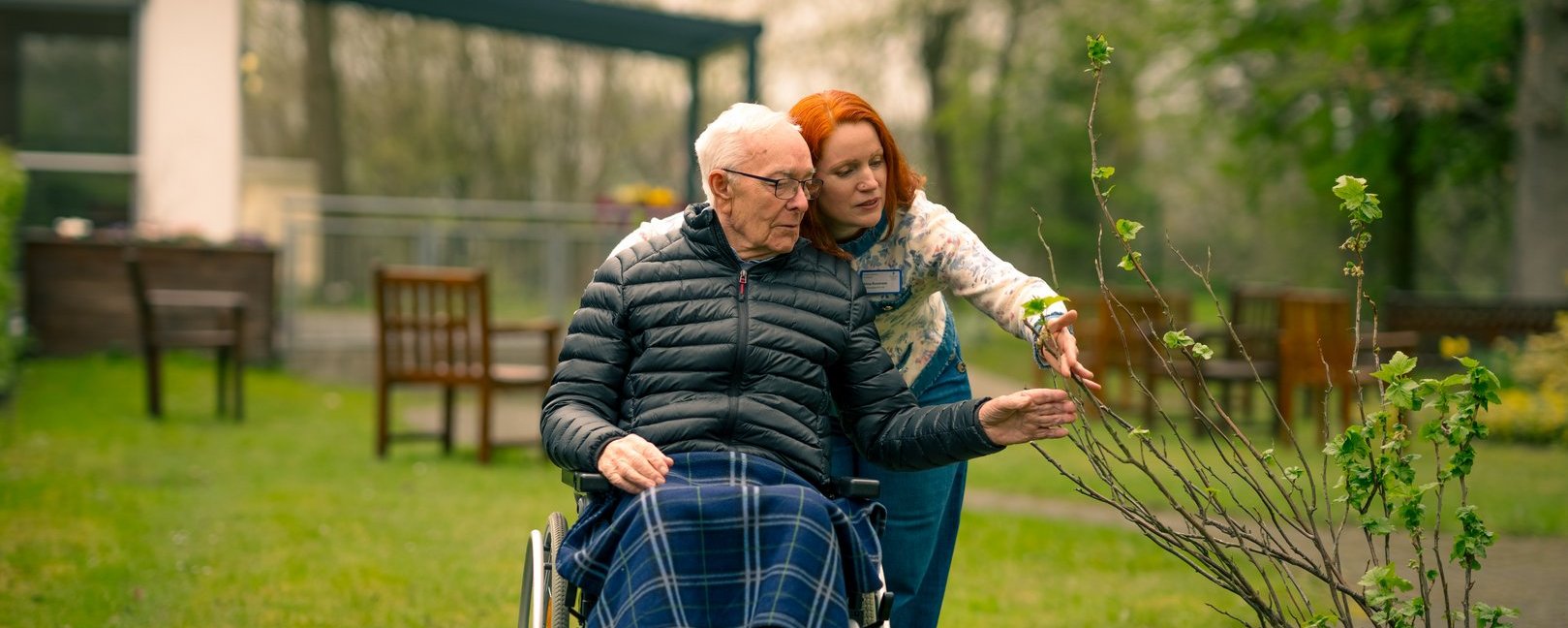 Ein älterer Mann im Rollstuhl wird von einer jungen Frau mit roten Haaren im Garten begleitet. Sie deutet auf eine junge Pflanze. Der Mann, der eine karierte Decke auf dem Schoß hat, betrachtet die Pflanze.