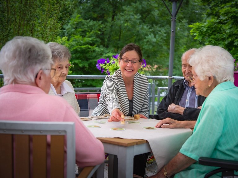 Vier Bewohner spielen mit einer Mitarbeiterin ein Brettspiel auf einer Terasse im Garten