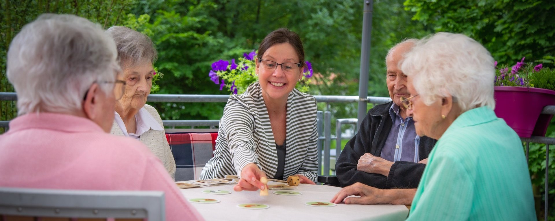 Vier Bewohner spielen mit einer Mitarbeiterin ein Brettspiel auf einer Terasse im Garten