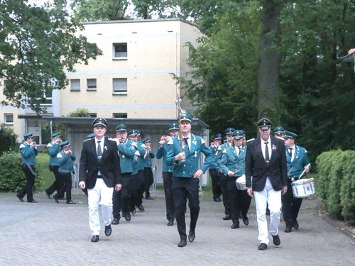 Eine Musikkapelle in grünen Uniformen marschiert auf einem Weg. Zwei Männer in schwarzen Jacken und weißen Hosen führen die Gruppe an, gefolgt von Musikern mit Flöten und Trommeln.