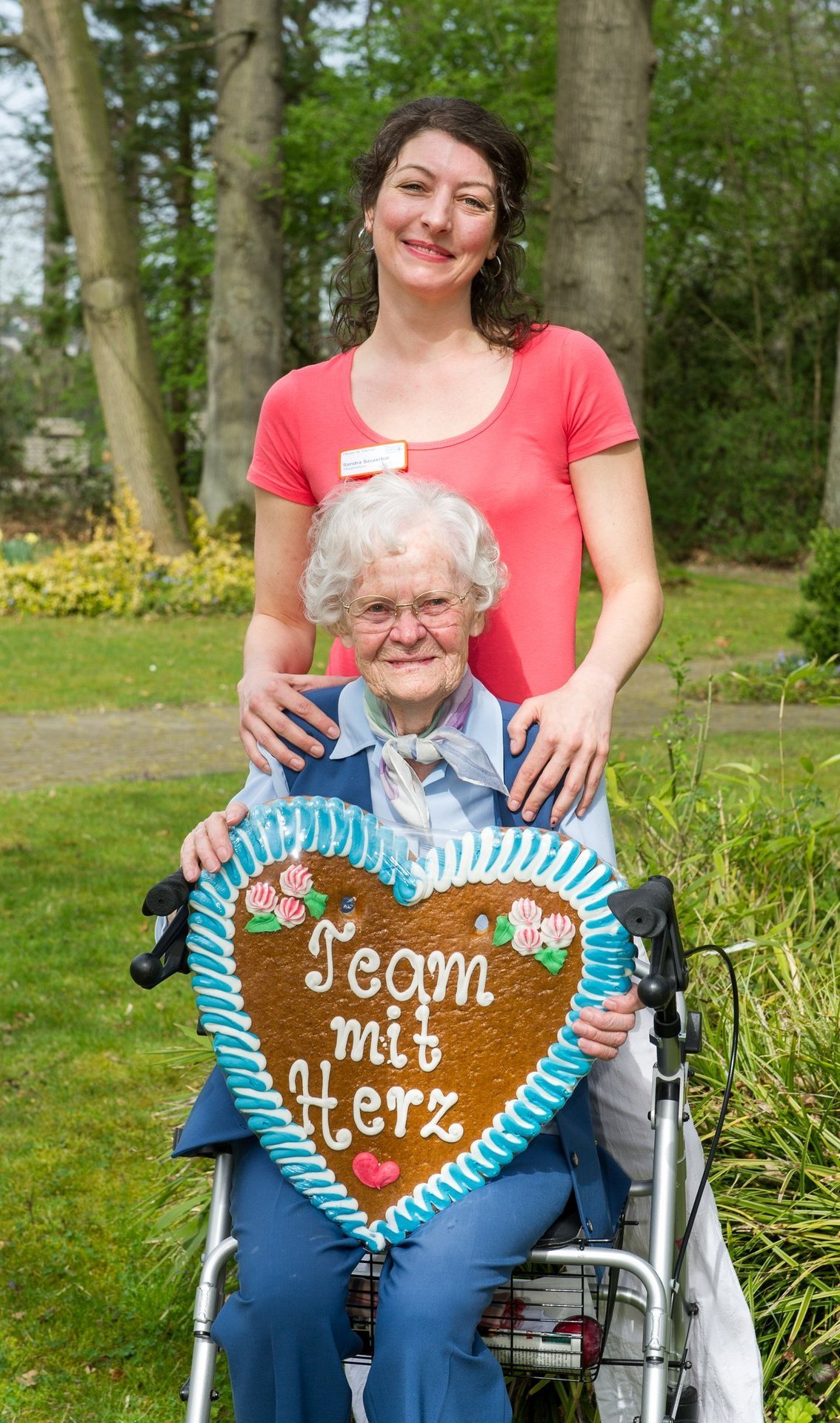 Eine junge Mitarbeiterin schiebt eine Bewohnerin in einem Rollstuhl mit einem großen Lebkuchenherz auf dem Schoß