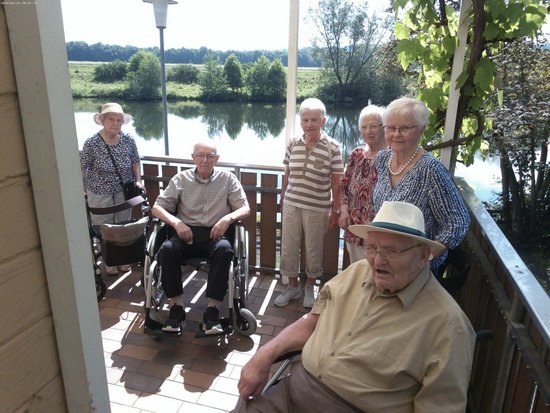 Sechs Senioren auf einer Holzterrasse mit Blick auf einen Fluss. Im Vordergrund sitzt ein Mann im Rollstuhl. Dahinter stehen vier Frauen und ein weiterer Mann im Rollstuhl.
