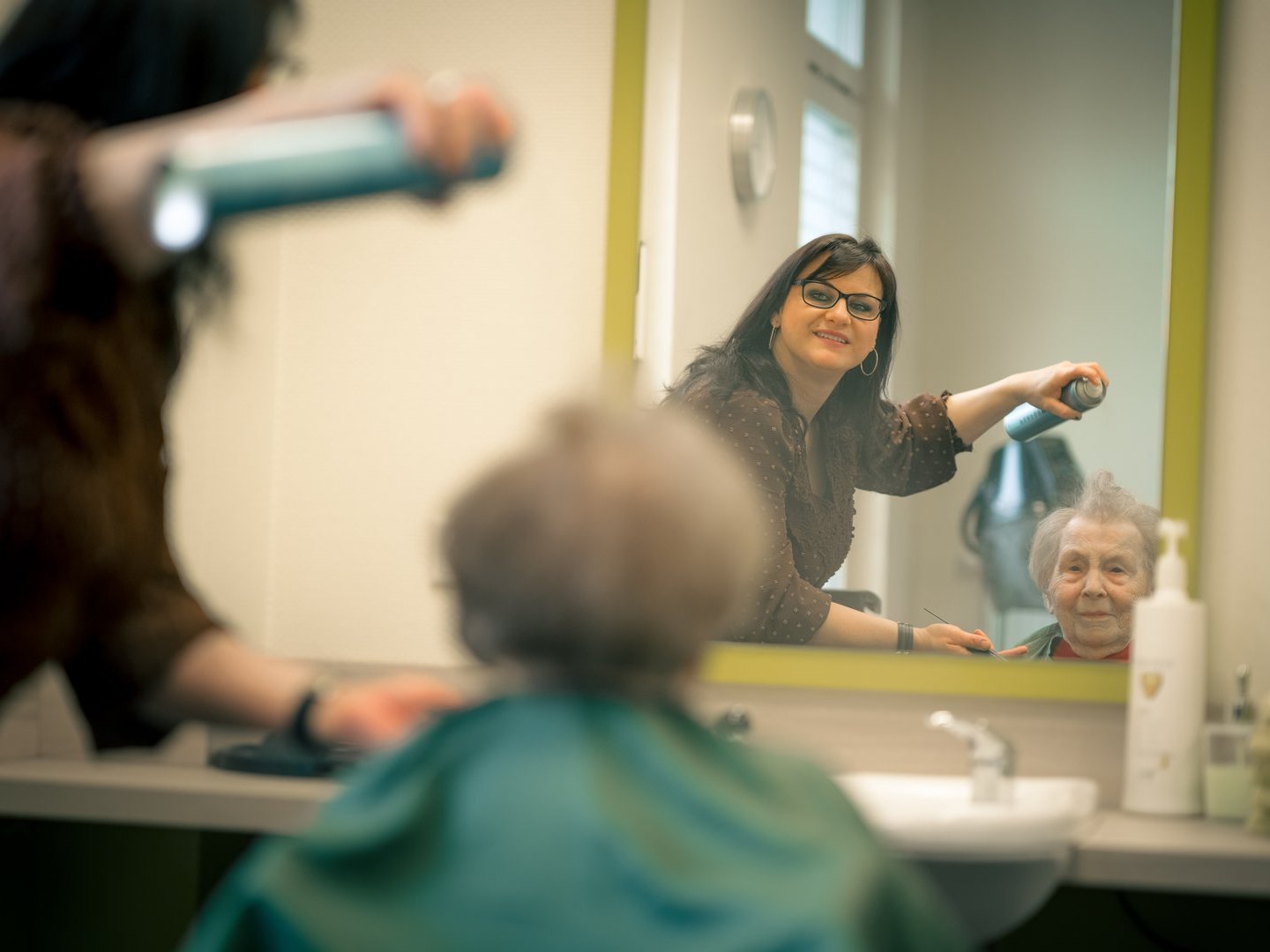 Eine Friseurin trocknet einer älteren Dame die Haare. Die Szene zeigt die Rückansicht der Kundin und im Spiegel das Lächeln der beiden Frauen sowie den Haartrockner in der Hand der Friseurin.