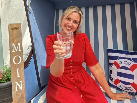 Eine lächelnde Frau in einem roten Kleid sitzt in einem blau-weiß gestreiften Strandkorb. Sie hält ein Wasserglas als Toast in die Kamera. Links steht ein Holzschild mit „MOIN", rechts liegt ein Kissen mit einem Rettungsring-Motiv.