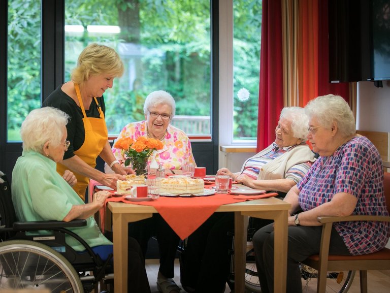 Vier Bewohner sitzen um einen Holztisch und bekommen von einer Mitarbeiterin Kuchen serviert.