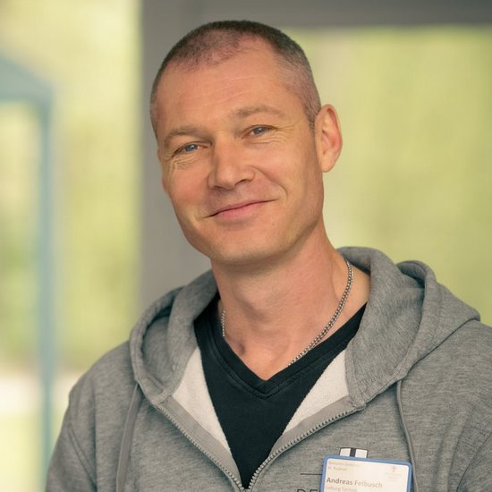 Andreas Felbusch Ein Porträt eines lächelnden Mannes mit kurzen Haaren, der einen grauen Kapuzenpullover über einem schwarzen T-Shirt trägt. An seiner Brust ist ein Namensschild mit der Aufschrift „DER ORDEN Wirtshaus“ zu sehen. Der Hintergrund ist unscharf und hell.