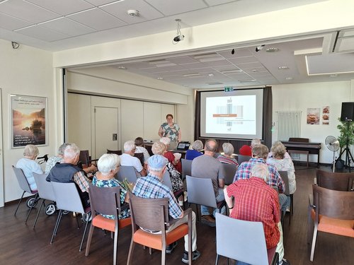 Eine Frau hält einen Vortrag vor einer Gruppe älterer Menschen. Sie steht neben einer Leinwand mit einer Präsentation, während das Publikum auf Stühlen sitzt und zuhört.