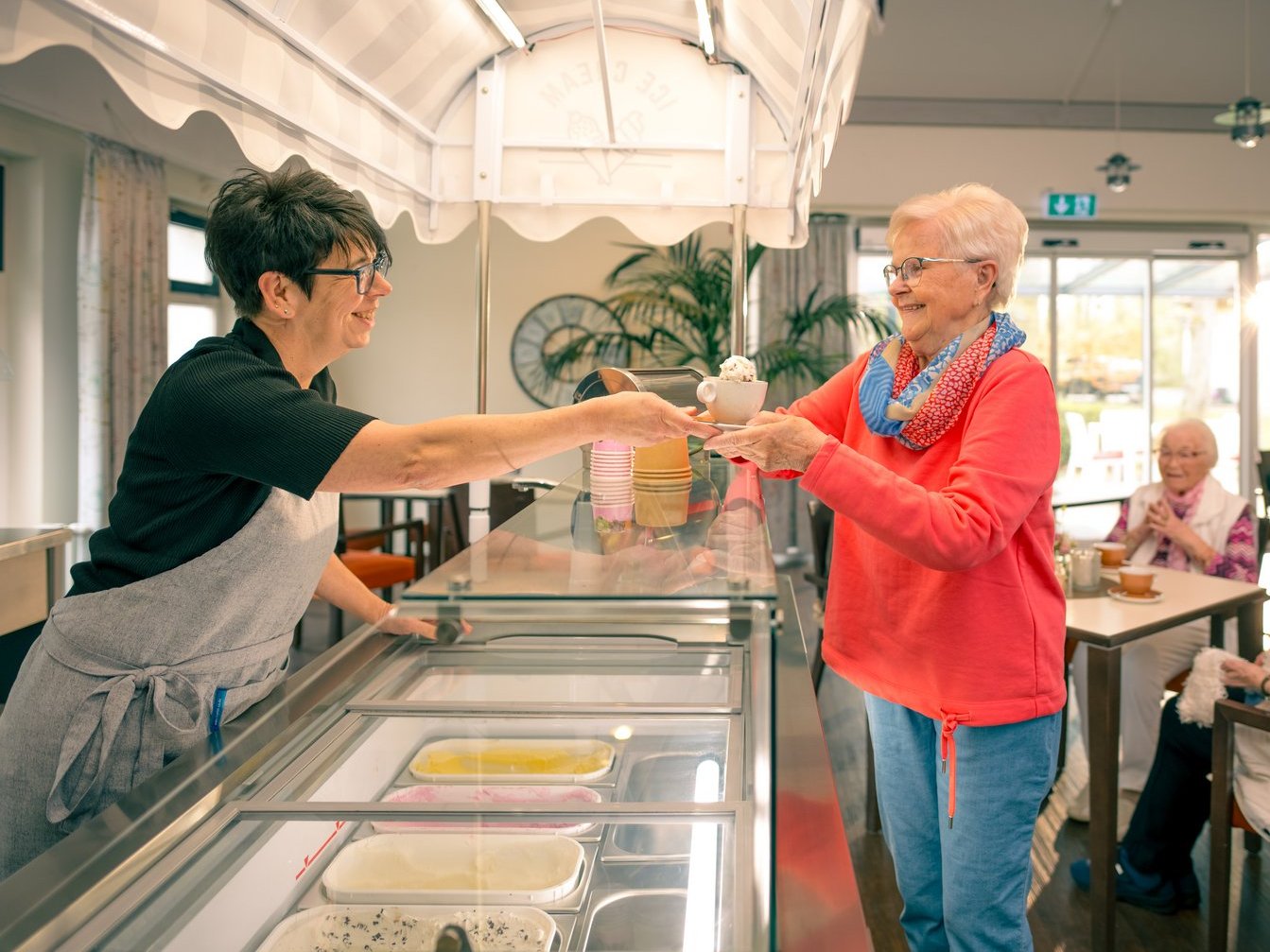 Eine freundliche Mitarbeiterin reicht einer Seniorin im roten Pullover ein Eis aus der Kühltheke. Beide lächeln sich an. Im Hintergrund sitzen weitere ältere Menschen im Raum.