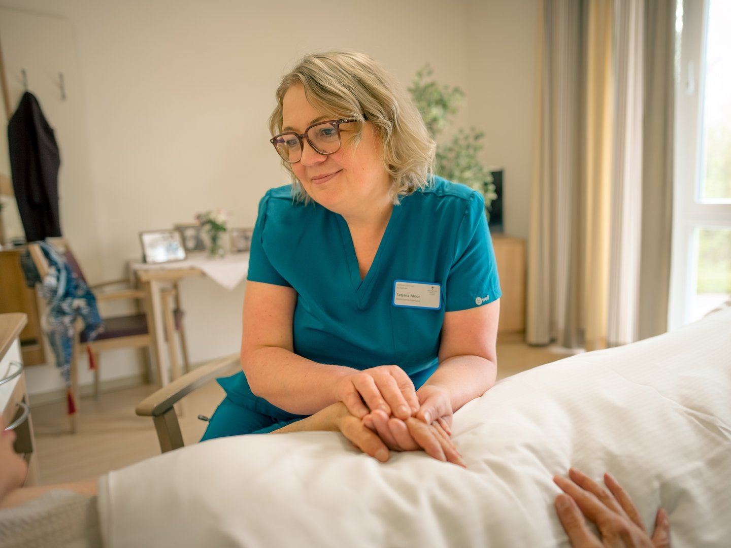 Eine Pflegekraft in blauer Kleidung sitzt am Bett eines Patienten und hält dessen Hand in einer tröstenden Geste, während sie ihn einfühlsam ansieht.